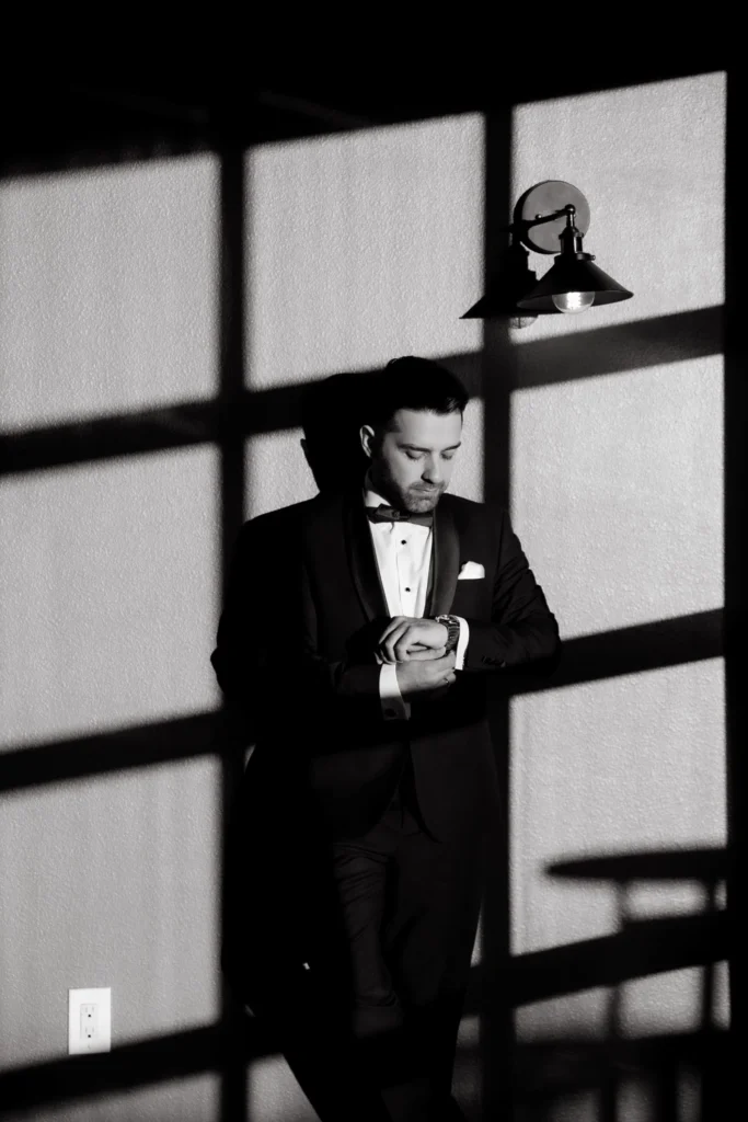 Black-and-white portrait of a groom adjusting his cuff in window light